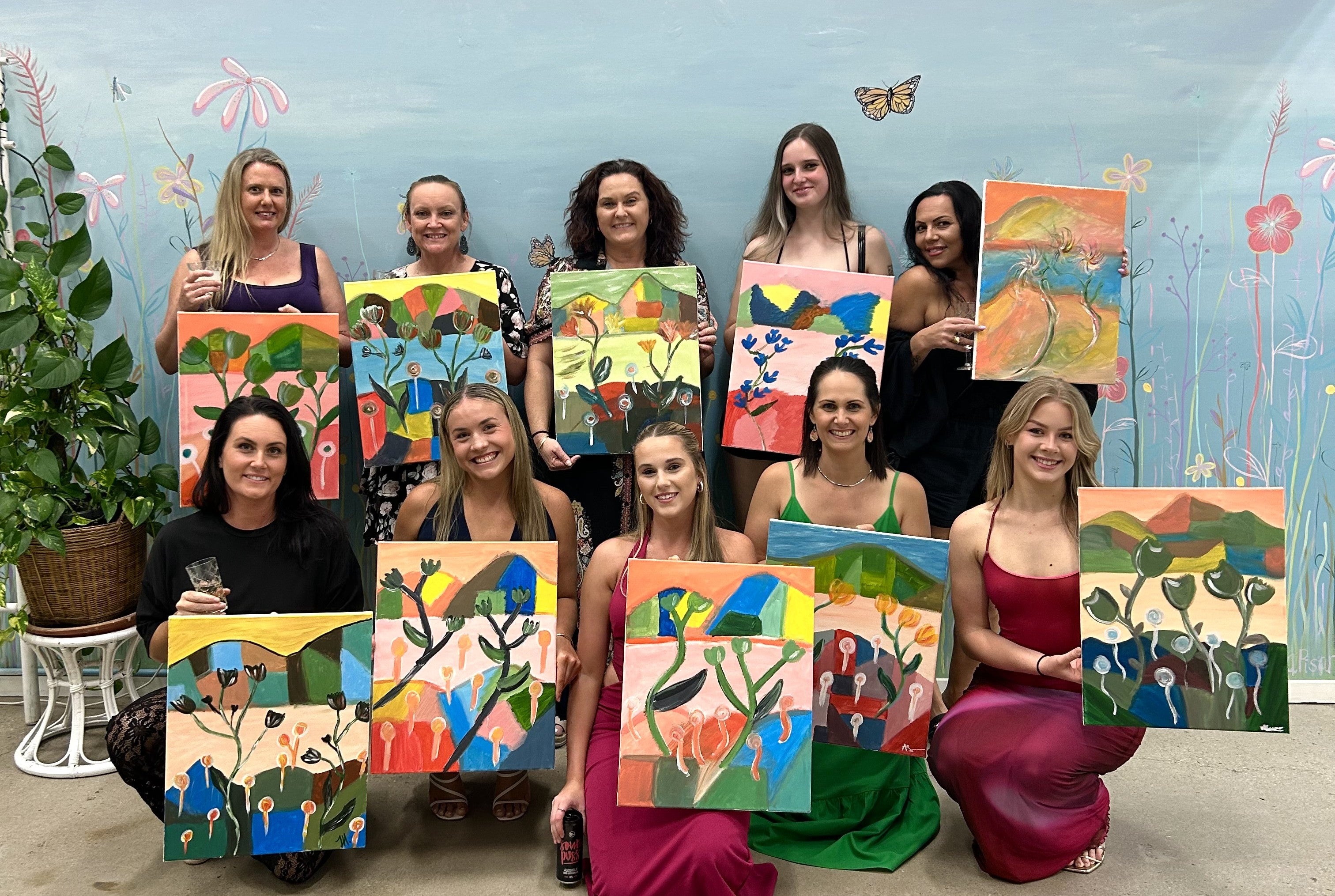 Group of women holding up their colorful paintings in a room with a butterfly decoration.