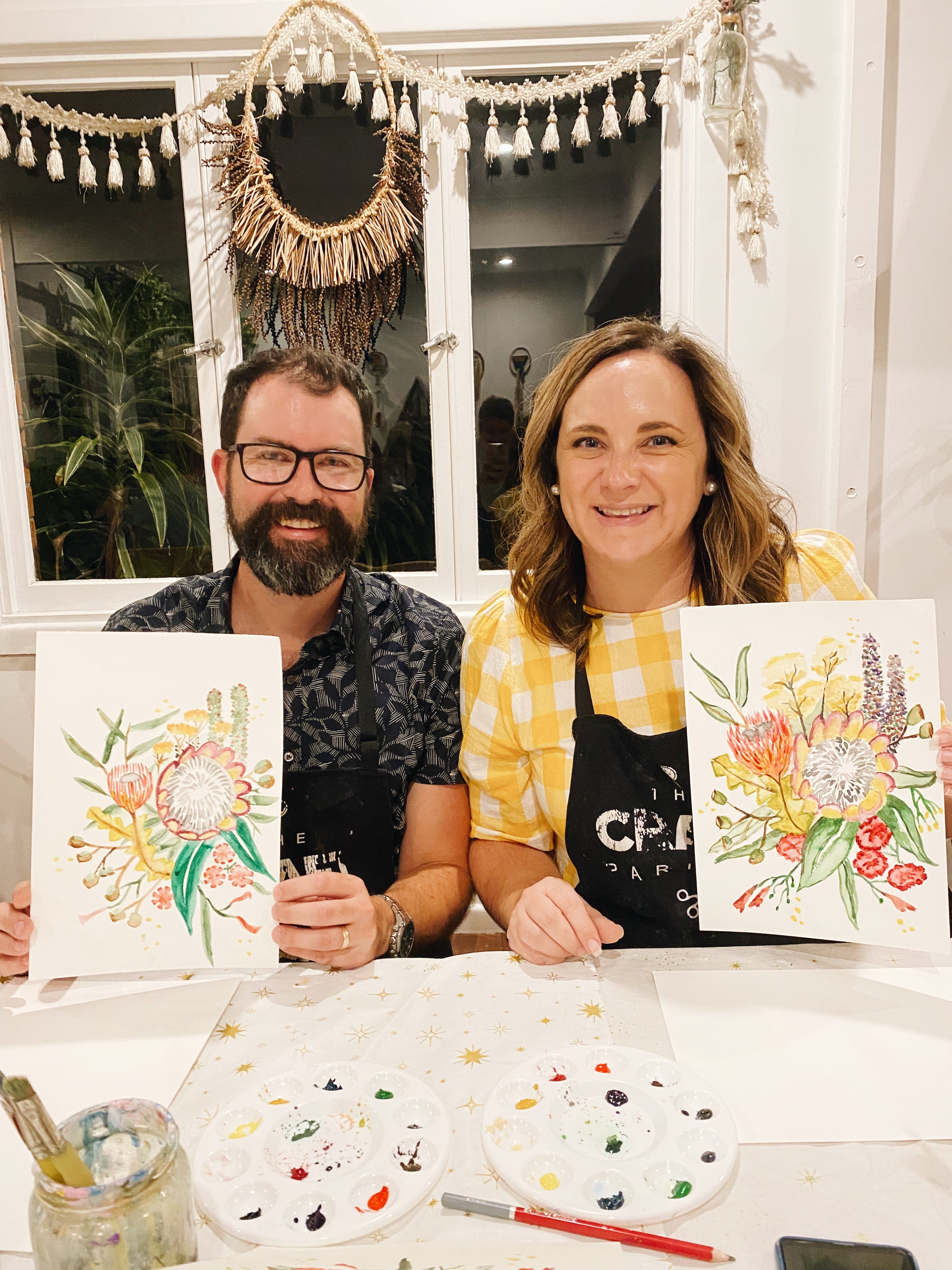 Two people holding floral paintings indoors with a decorative window in the background.