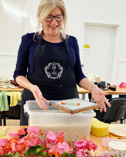 Woman making paper, standing behind a table with flowers and a container.