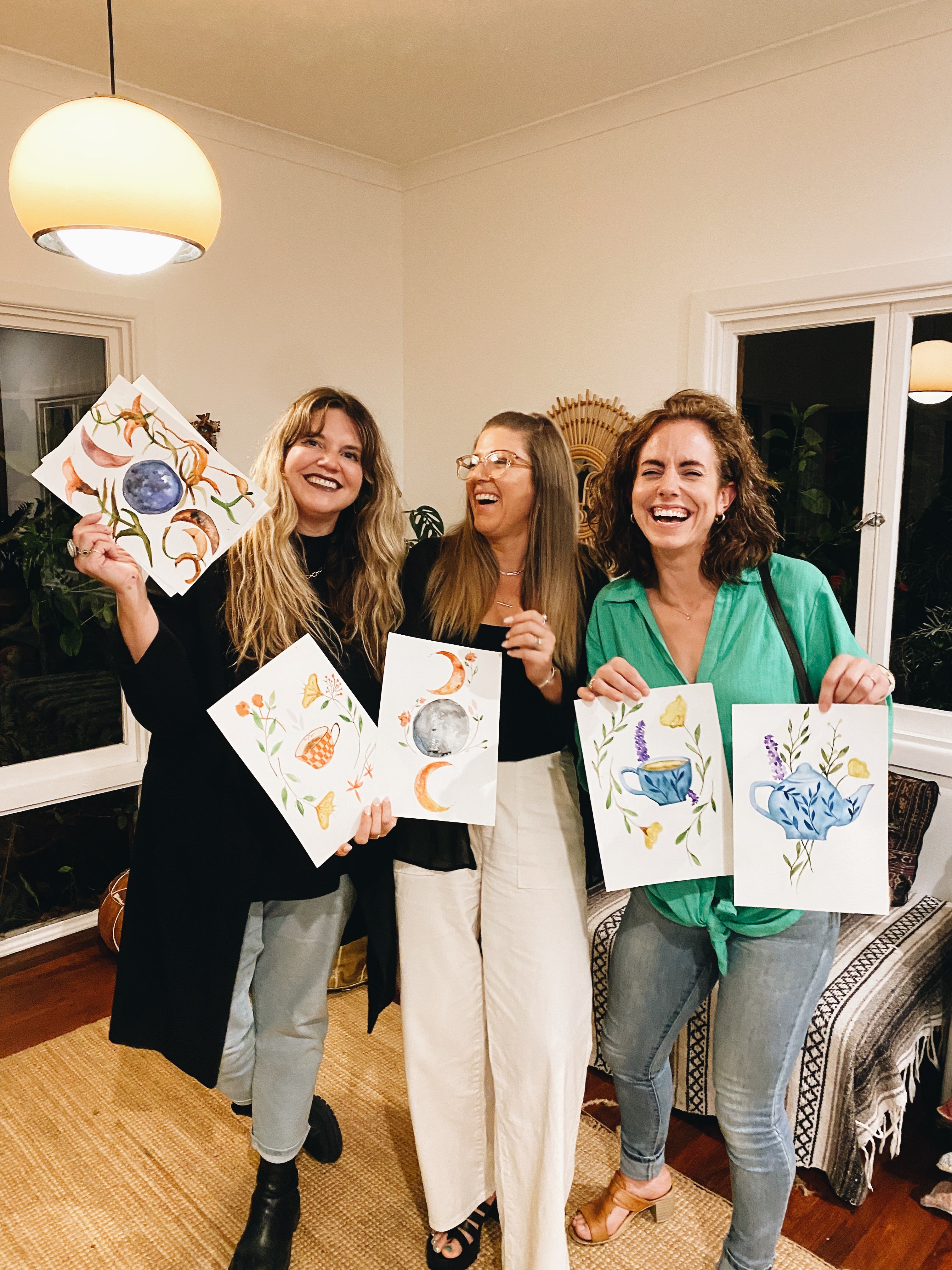 Three women holding up their artwork in a home setting