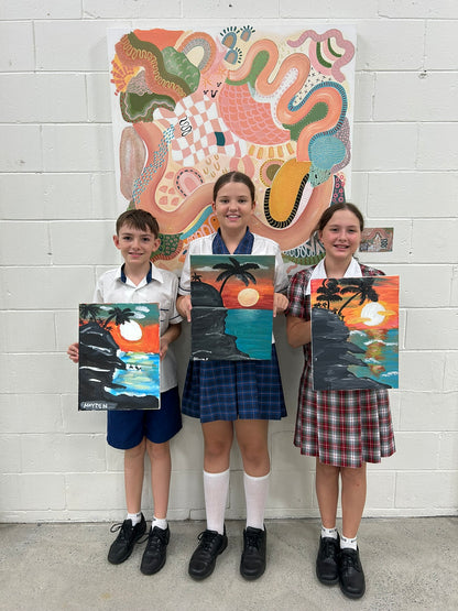 Three children holding artworks in front of a large colorful painting on a white brick wall.