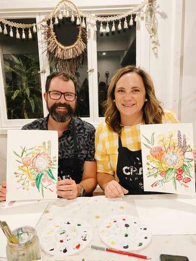 Two people holding floral paintings indoors with a decorative window in the background.