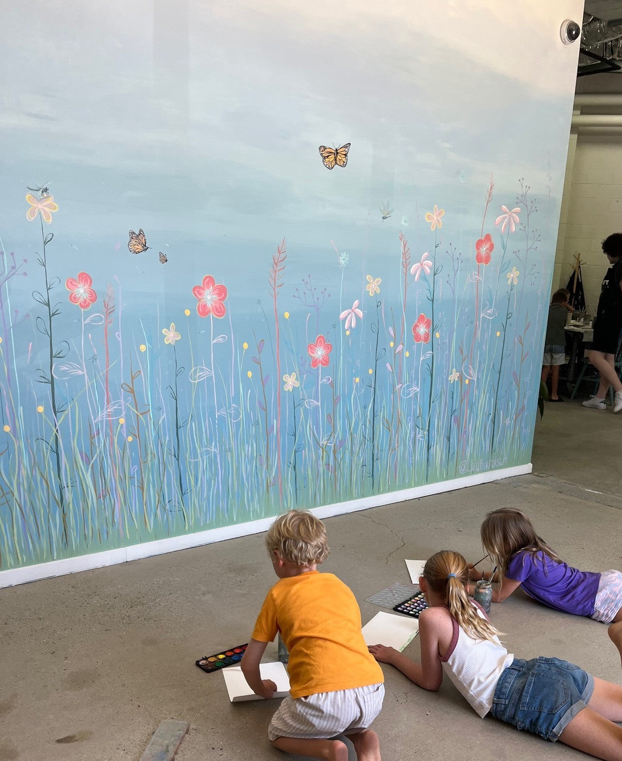 Children sitting on the floor in front of a large mural of flowers and butterflies.