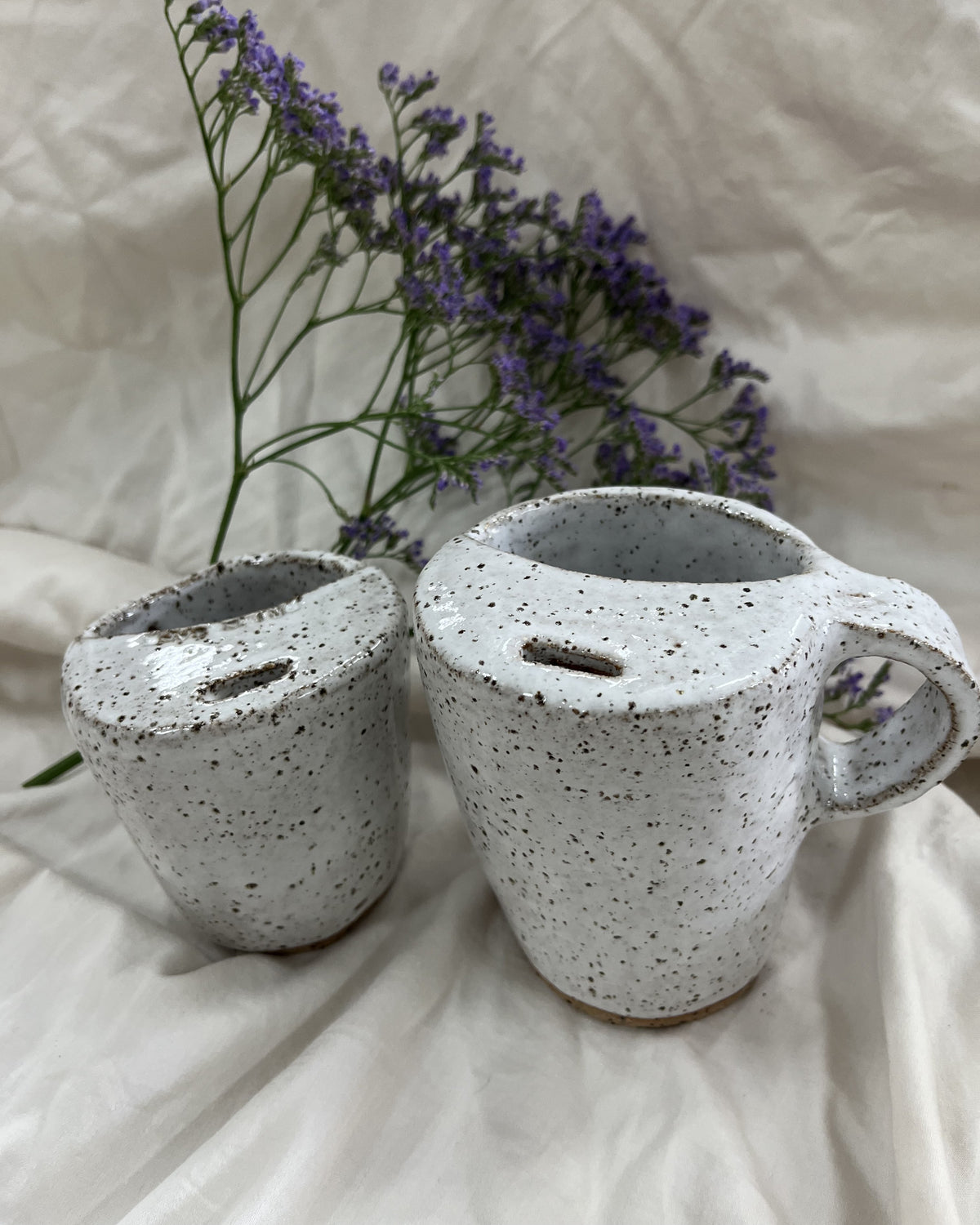 Two speckled ceramic mugs in a light gray-white glaze with dark brown/black flecks, displayed on a draped white fabric backdrop with purple flowers in the background.
