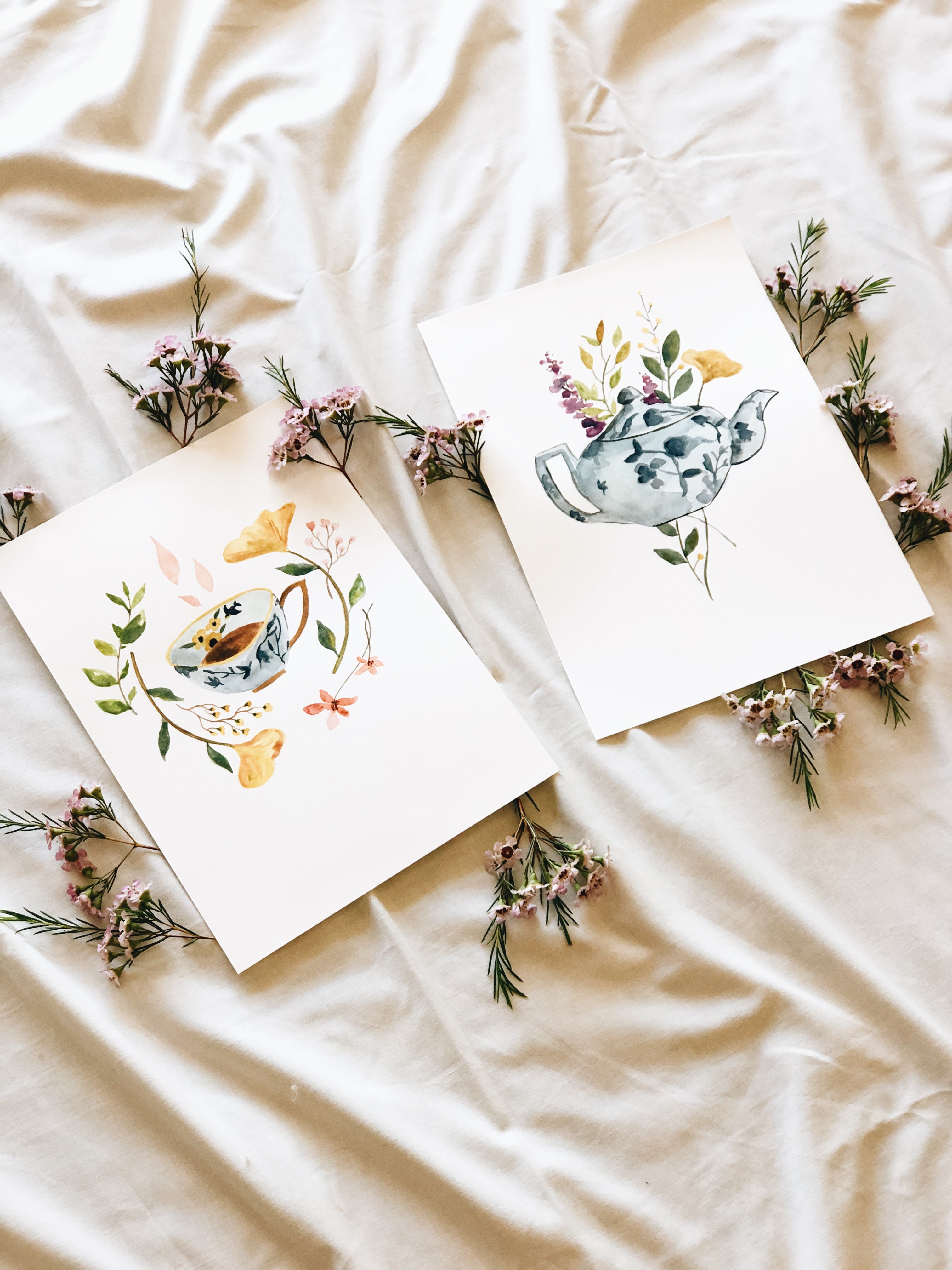 Two cards with teapot designs and floral elements on a beige fabric background.