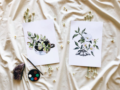 Two botanical prints on a textured fabric surface with paint supplies.